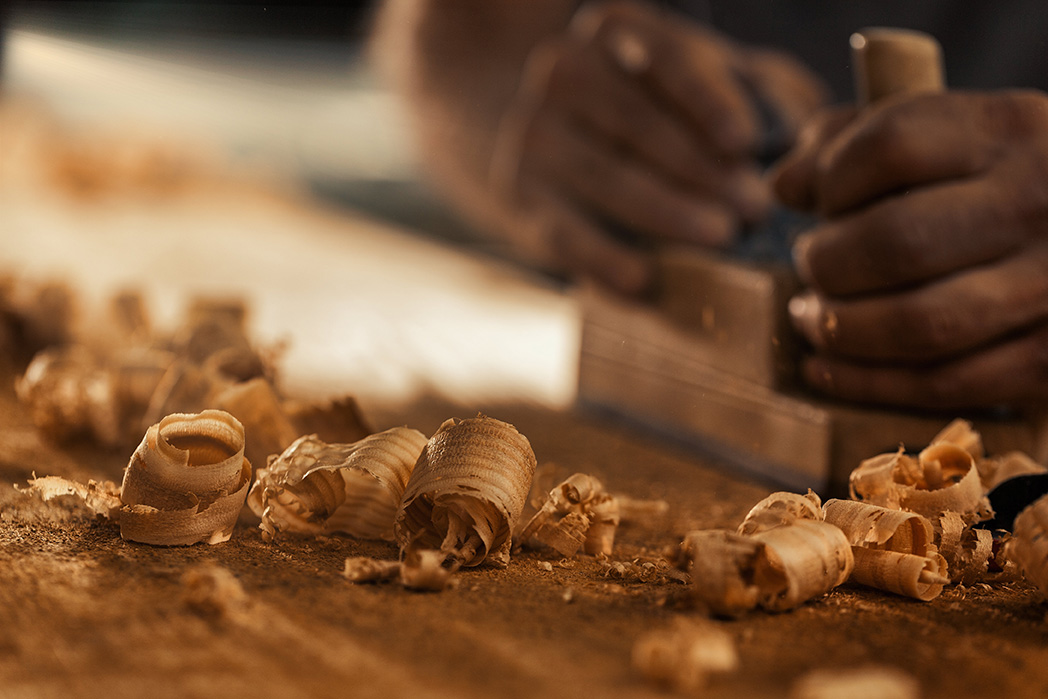The wood artisan, a robust and strong man known as a carpenter, hand planes a board in the traditional manner, producing shavings of sawdust that look like golden curls of wood. It is a strenuous and